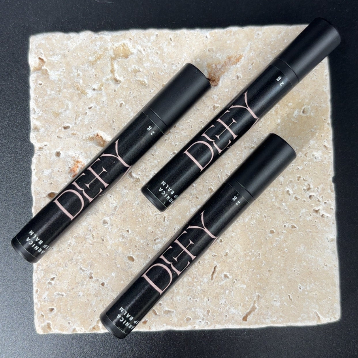Four black lip products on a beige stone surface with a black background