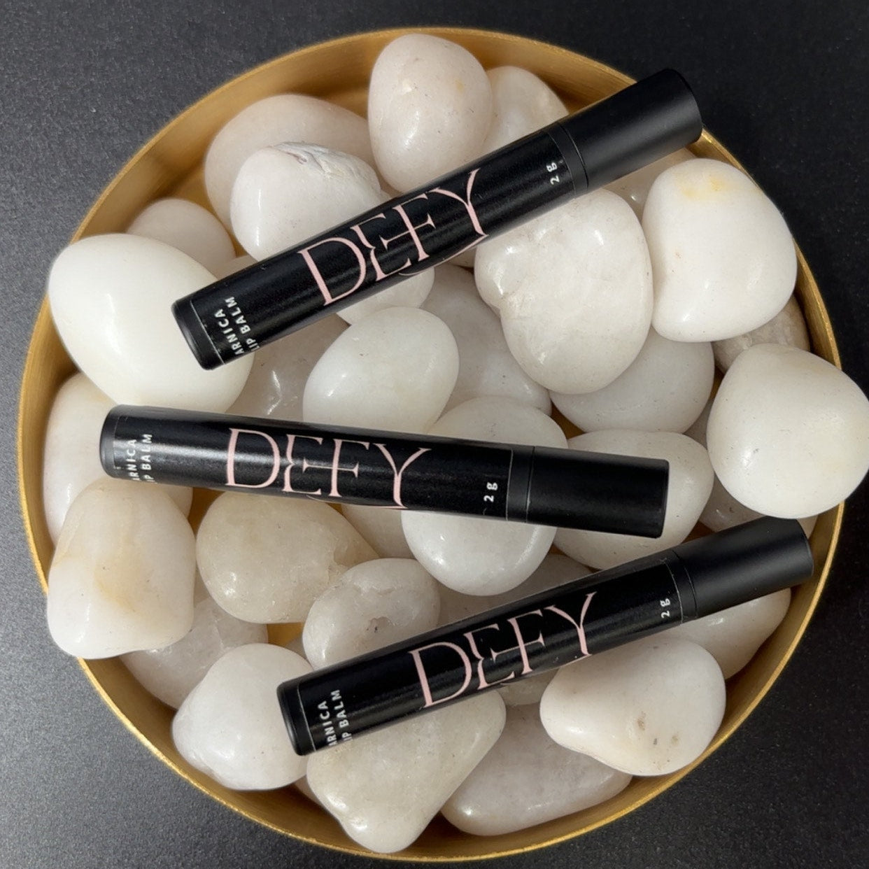 Four black makeup products with Arnica Lip balm and Defy branding on a bed of white stones.